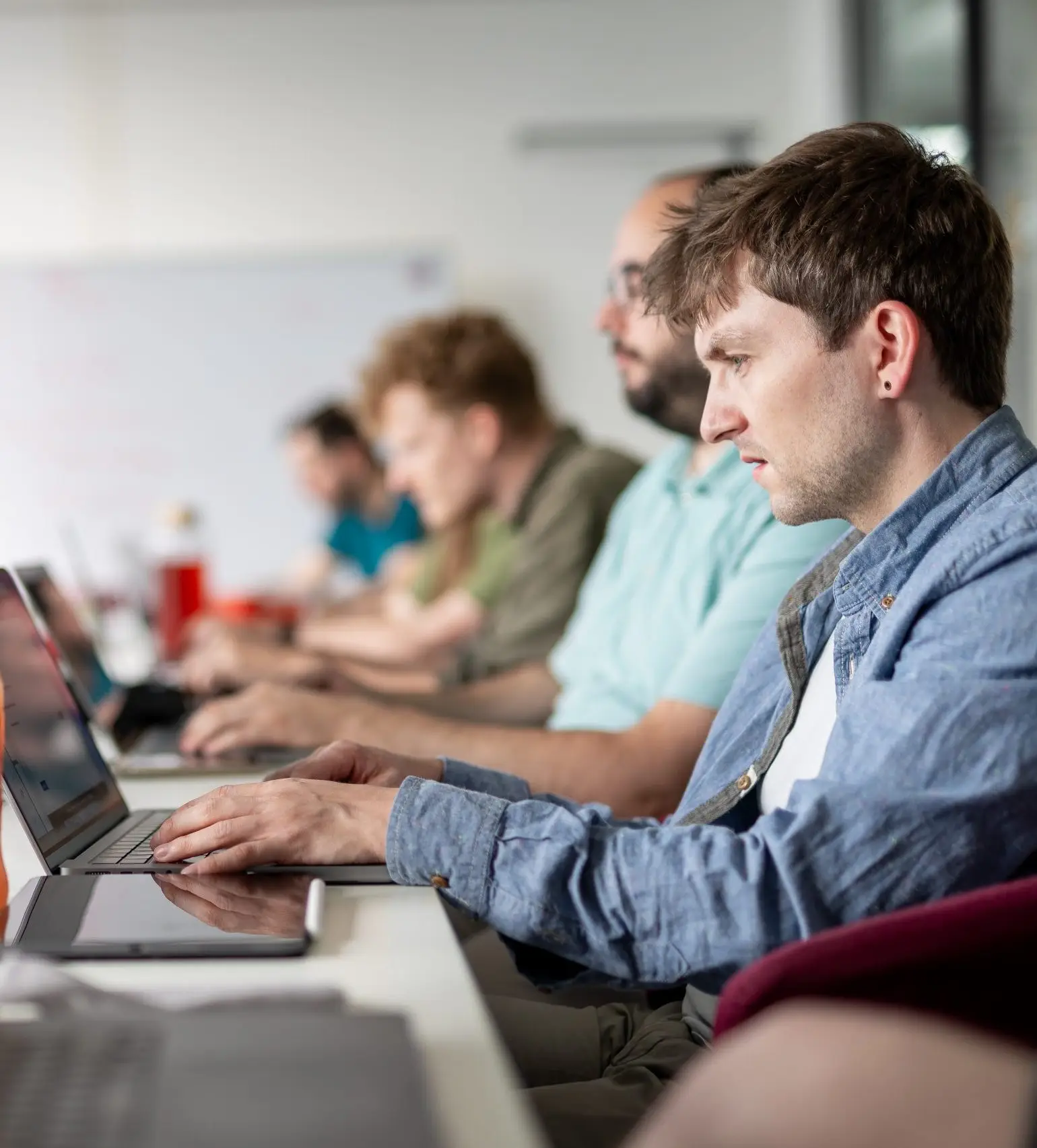 Eine Gruppe von Menschen sitzt an einem langen Tisch in einem hellen Büro und arbeitet konzentriert an Laptops. Im Vordergrund ist eine Flasche Getränk und ein Glas Wasser zu sehen. Im Hintergrund steht eine Tafel mit Notizen, während die Atmosphäre auf Teamarbeit und Fokus hinweist