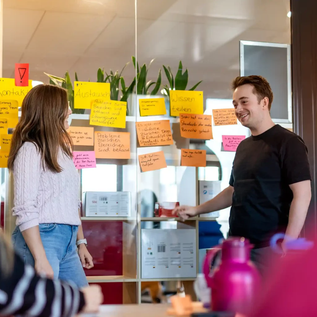 Zwei Personen im Gespräch vor einer Glaswand mit bunten Post-its in einem Büro. Die Wand ist mit handschriftlichen Notizen bedeckt, im Hintergrund sind Pflanzen und Möbel sichtbar.