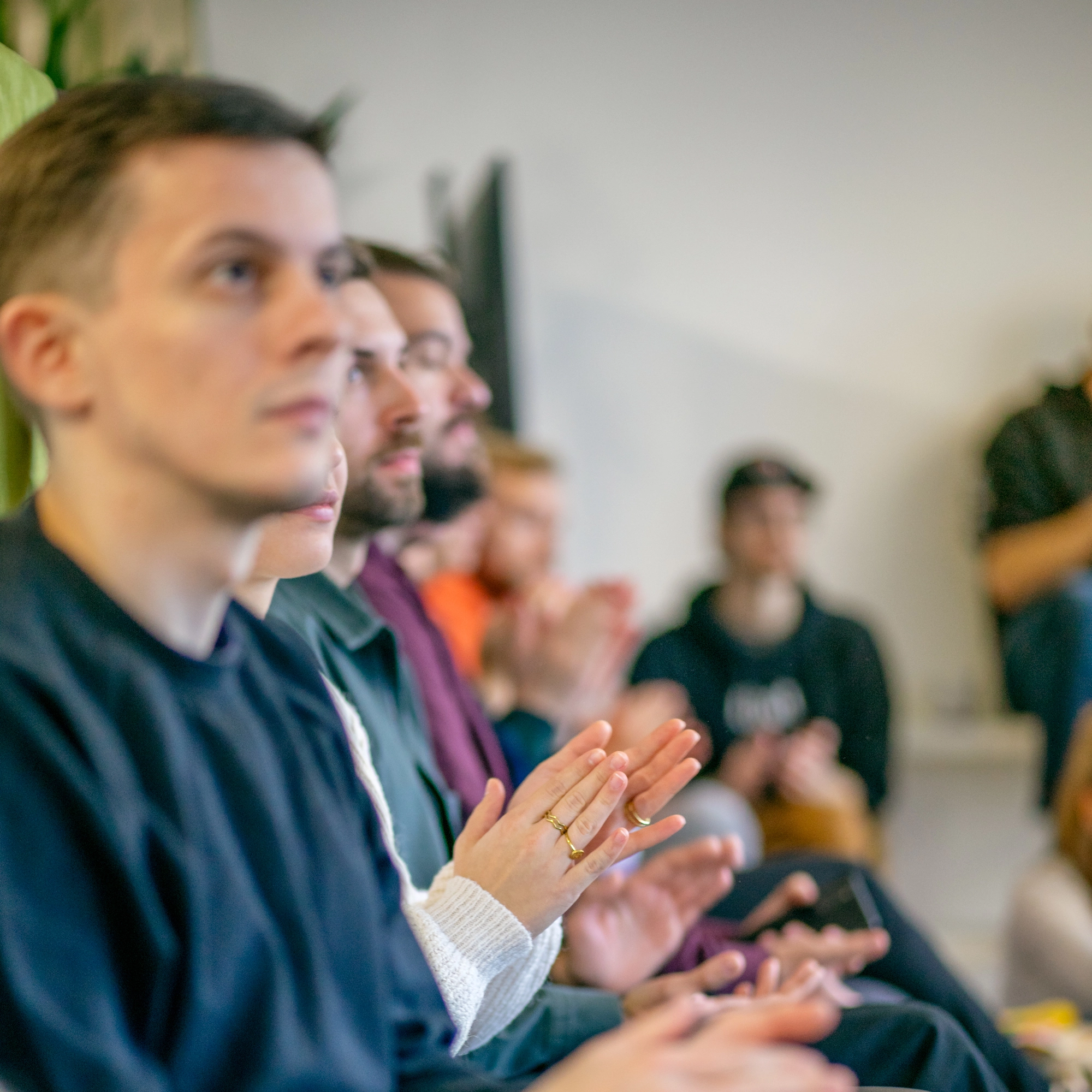 Eine Gruppe von Menschen sitzt in einem Raum und applaudiert. Der Fokus liegt auf den vorderen Reihen, wo Personen in Alltagskleidung klatschen. Der Hintergrund ist leicht unscharf, zeigt jedoch weitere Teilnehmer und eine informelle Atmosphäre.