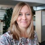 Ansprechpartnerin für Workshops in einem Büro, mit schulterlangem Haar und gemustertem Oberteil. Im Hintergrund sind eine Pflanze und Büromöbel sichtbar.