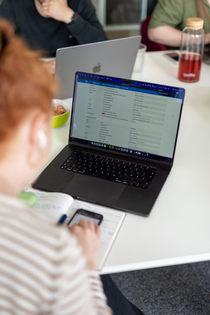 Person arbeitet mit Laptop und Smartphone an einem Tisch, auf dem auch ein Notizbuch, eine grüne Schale und eine Flasche mit roter Flüssigkeit stehen. Im Hintergrund sitzt eine weitere Person mit Laptop.