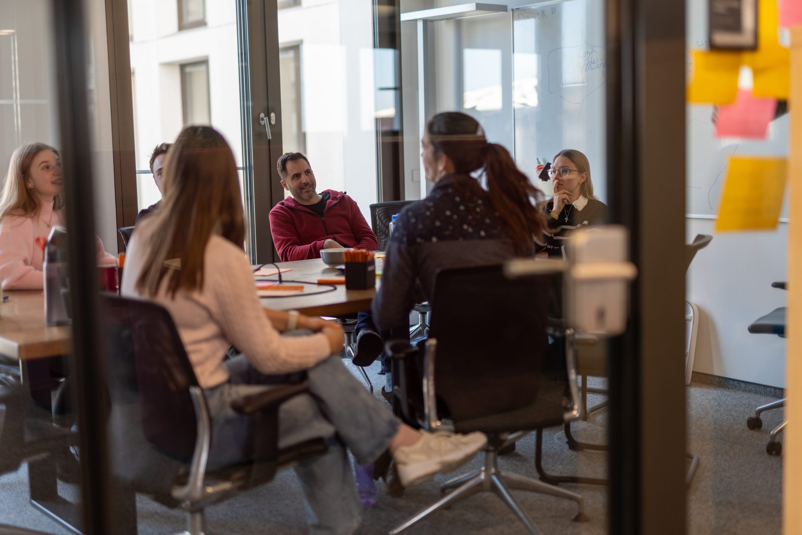 Teammeeting in einem modernen Konferenzraum mit sechs Personen, die gemeinsam an einem Tisch sitzen und sich beraten. Im Vordergrund sind Post-its an einer Glastür sichtbar.