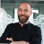 Ansprechpartner für Wartung und Service in einem Büro mit gefrosteten Glaswänden mit Kreismuster, trägt ein schwarzes Shirt und hat einen Bart.