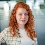 Ansprechpartnerin für UX Design in einem modernen Büro, mit lockigem roten Haar und kariertem Hemd über weißem Oberteil, vor einer Glaswand mit unscharfem Hintergrund.