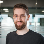 Ansprechpartner für Evaluierung und Testing in einem modernen Büro, mit Brille und Bart, trägt ein dunkles Shirt. Im Hintergrund sind Glaswände und unscharfe Büroelemente sichtbar.