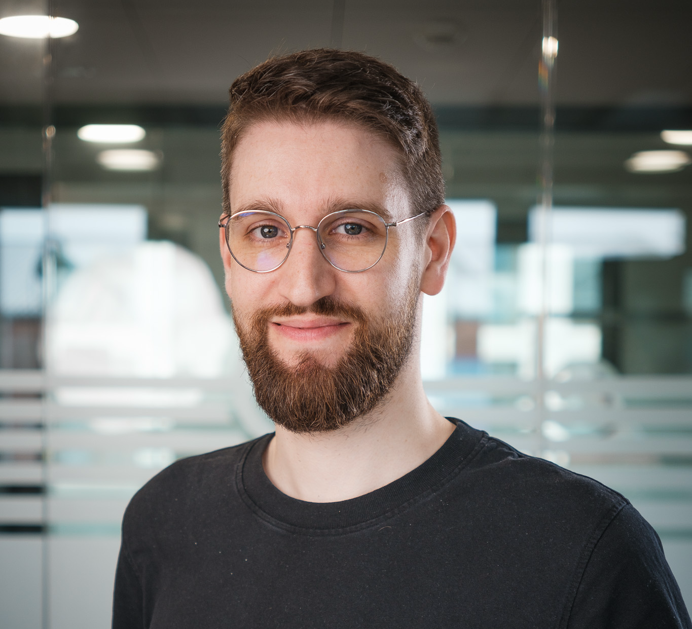 Ansprechpartner für Evaluierung und Testing in einem modernen Büro, mit Brille und Bart, trägt ein dunkles Shirt. Im Hintergrund sind Glaswände und unscharfe Büroelemente sichtbar.