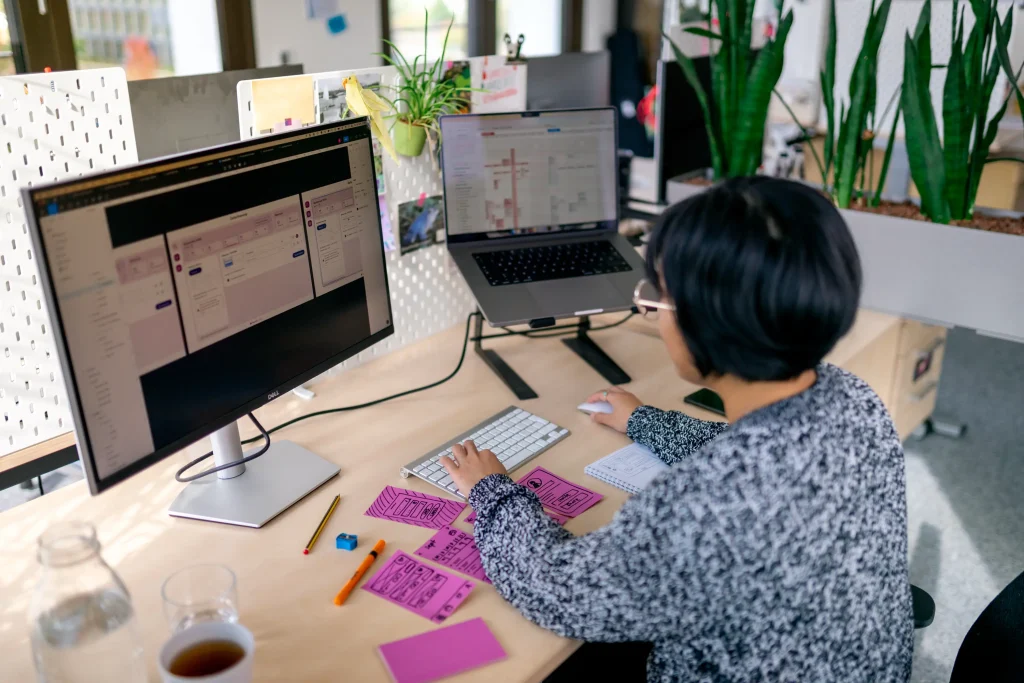 Person arbeitet an einem Schreibtisch mit zwei Monitoren, Tastatur und Maus. Auf dem Tisch liegen Post-its, im Hintergrund sind Pflanzen sichtbar. Die Bildschirme zeigen arbeitsbezogene Inhalte.
