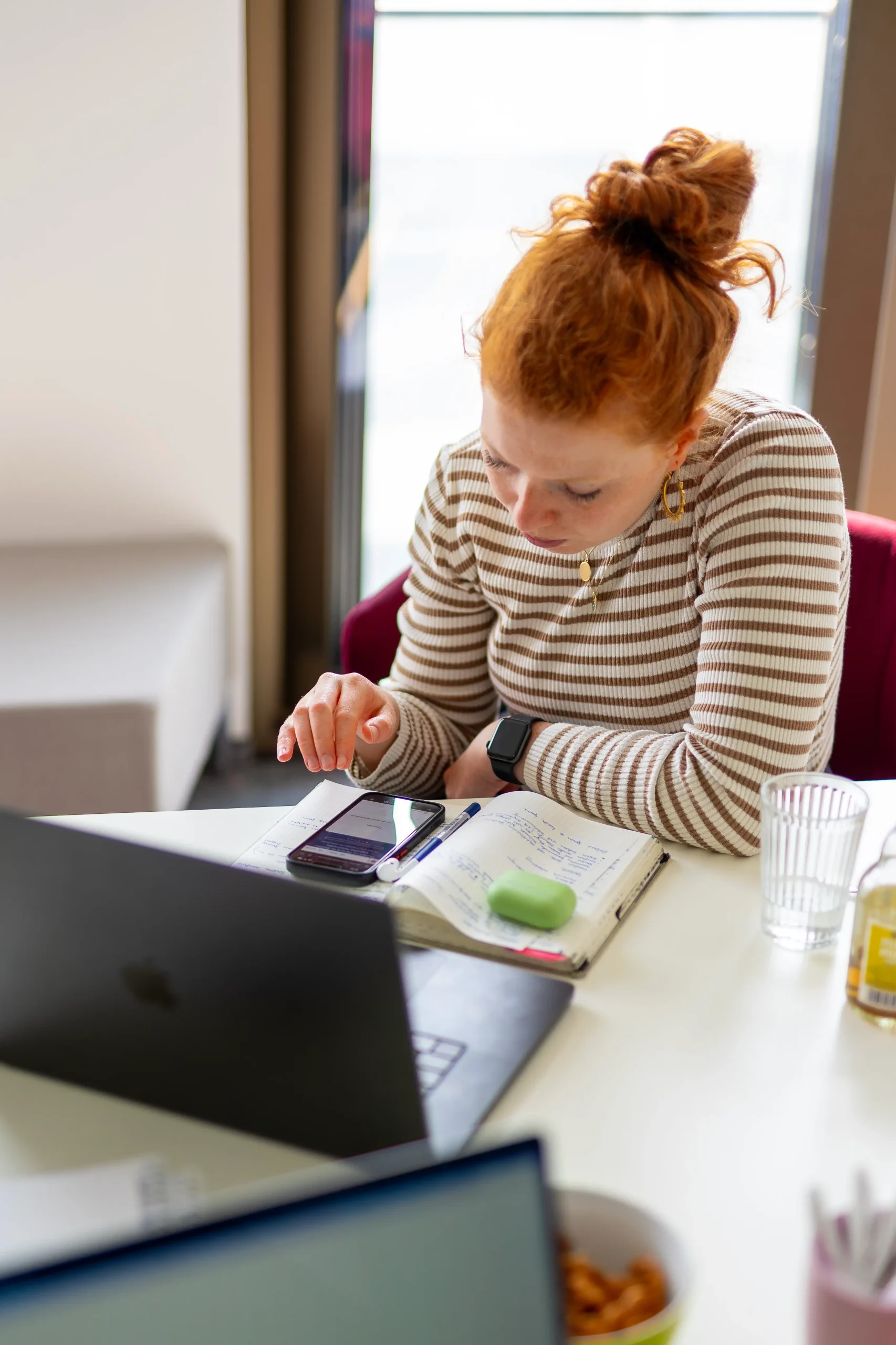 Person sitzt an einem Schreibtisch mit Laptop, Smartphone und offenem Notizbuch. Auf dem Tisch stehen ein Glas Wasser und Schreibutensilien, im Hintergrund fällt Tageslicht durch ein Fenster.