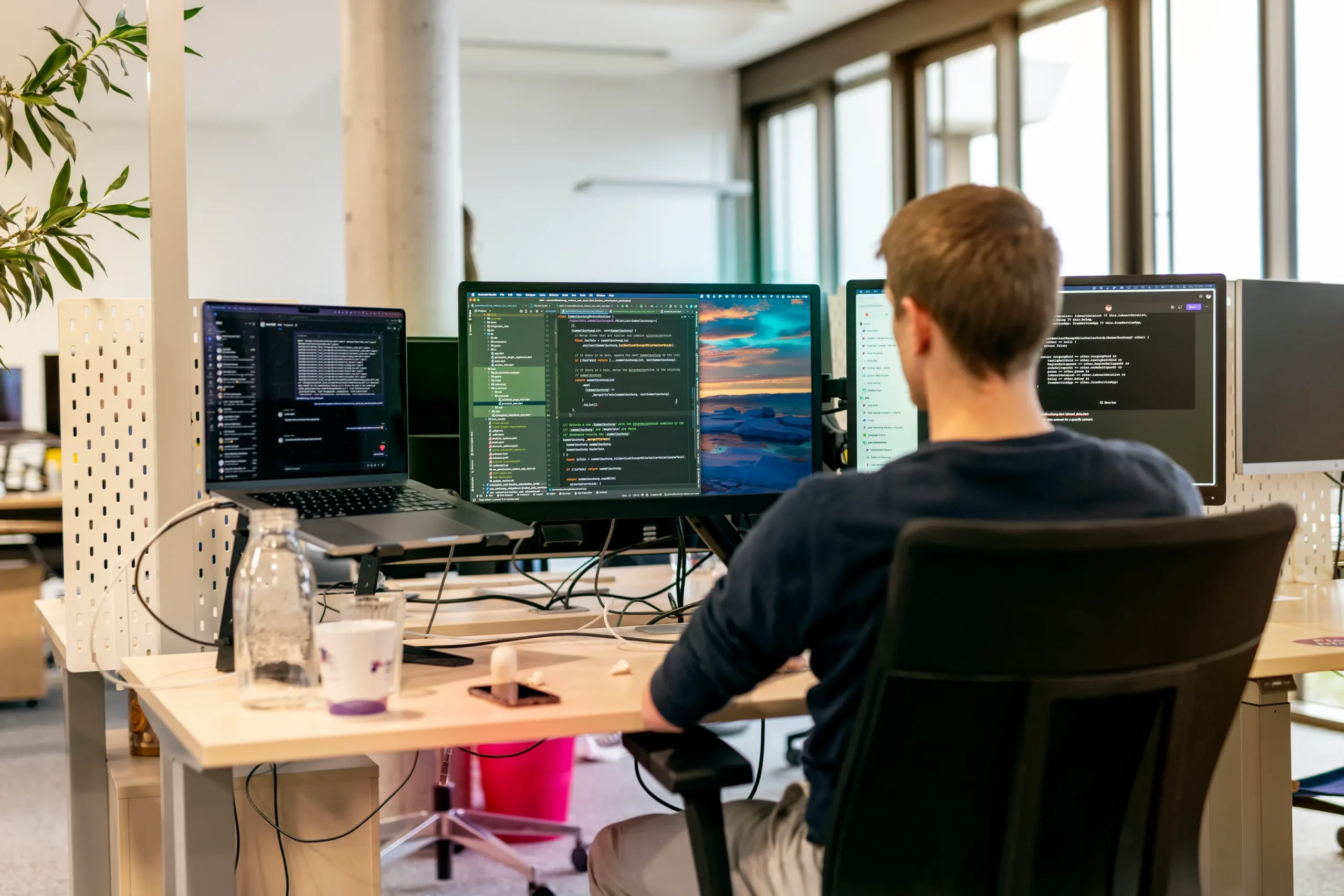 Person arbeitet in einem modernen Büro an vier Monitoren mit geöffneten Code-Editoren und Terminalfenstern. Auf dem Schreibtisch stehen eine Wasserflasche, eine Kaffeetasse und Unterlagen.