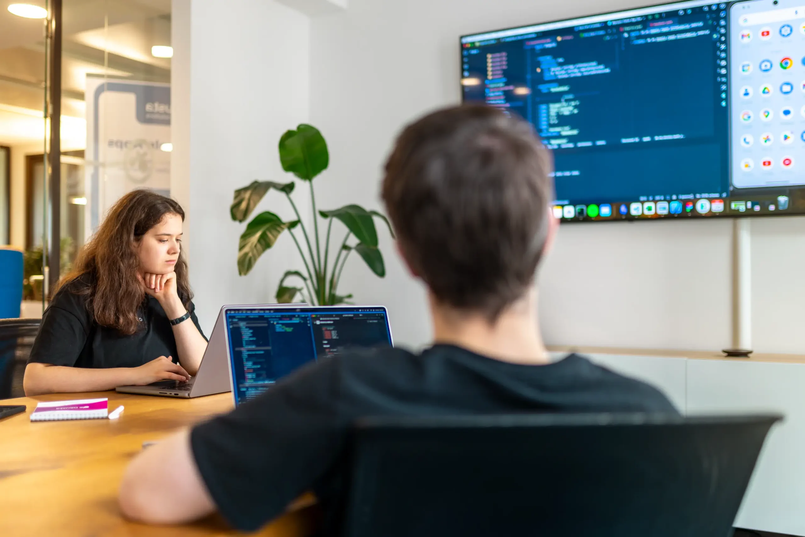 Zwei Personen arbeiten in einem modernen Büro an Code-Projekten. Eine Person sitzt am Laptop, die andere blickt auf einen großen Monitor mit Code und App-Icons. Im Hintergrund sind Pflanzen sichtbar.